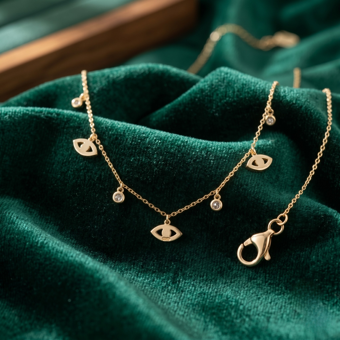 A delicate gold chain necklace with three small eye charms and tiny clear stones, displayed on draped emerald green velvet fabric.