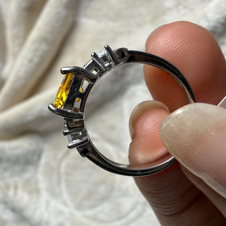 A close-up of a silver ring with a yellow gemstone held between fingers against a soft white fabric background.