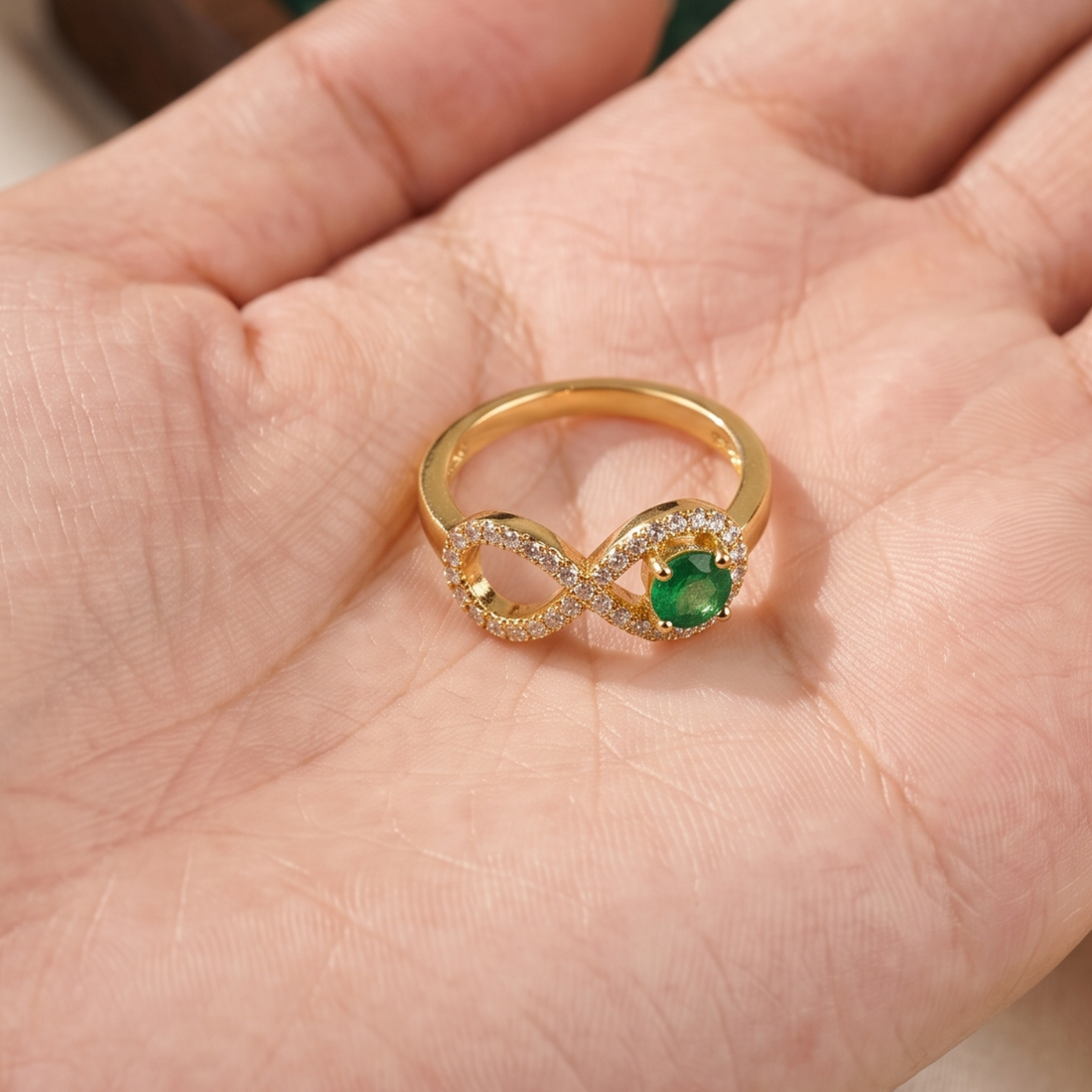 A gold infinity ring with a green emerald-cut center stone and pavé-set clear accent stones, resting on a person's palm.