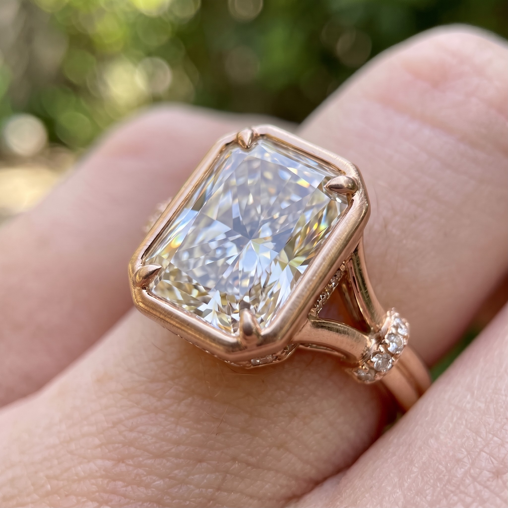 A close-up of a rose gold ring with a large rectangular clear gemstone being worn on a finger, with a blurred green outdoor background.