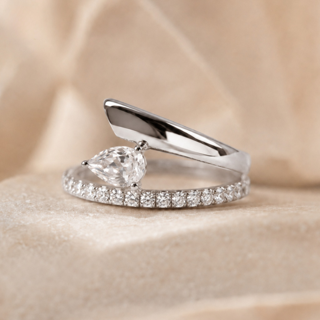 A silver-tone ring featuring a pear-cut clear gemstone and a pavé band, displayed on a soft beige fabric background.