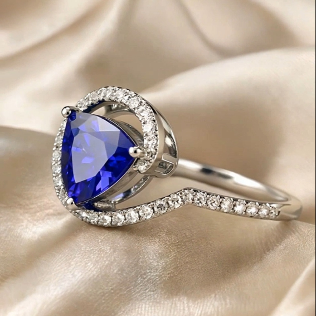 A silver ring featuring a large pear-cut deep blue gemstone surrounded by a halo of small clear stones, with a wavy band also set with clear stones, resting on draped champagne-colored satin fabric.