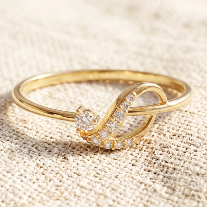 A gold ring with a stylized infinity symbol design, featuring a round clear gemstone and a row of small clear stones, resting on a textured beige fabric background.