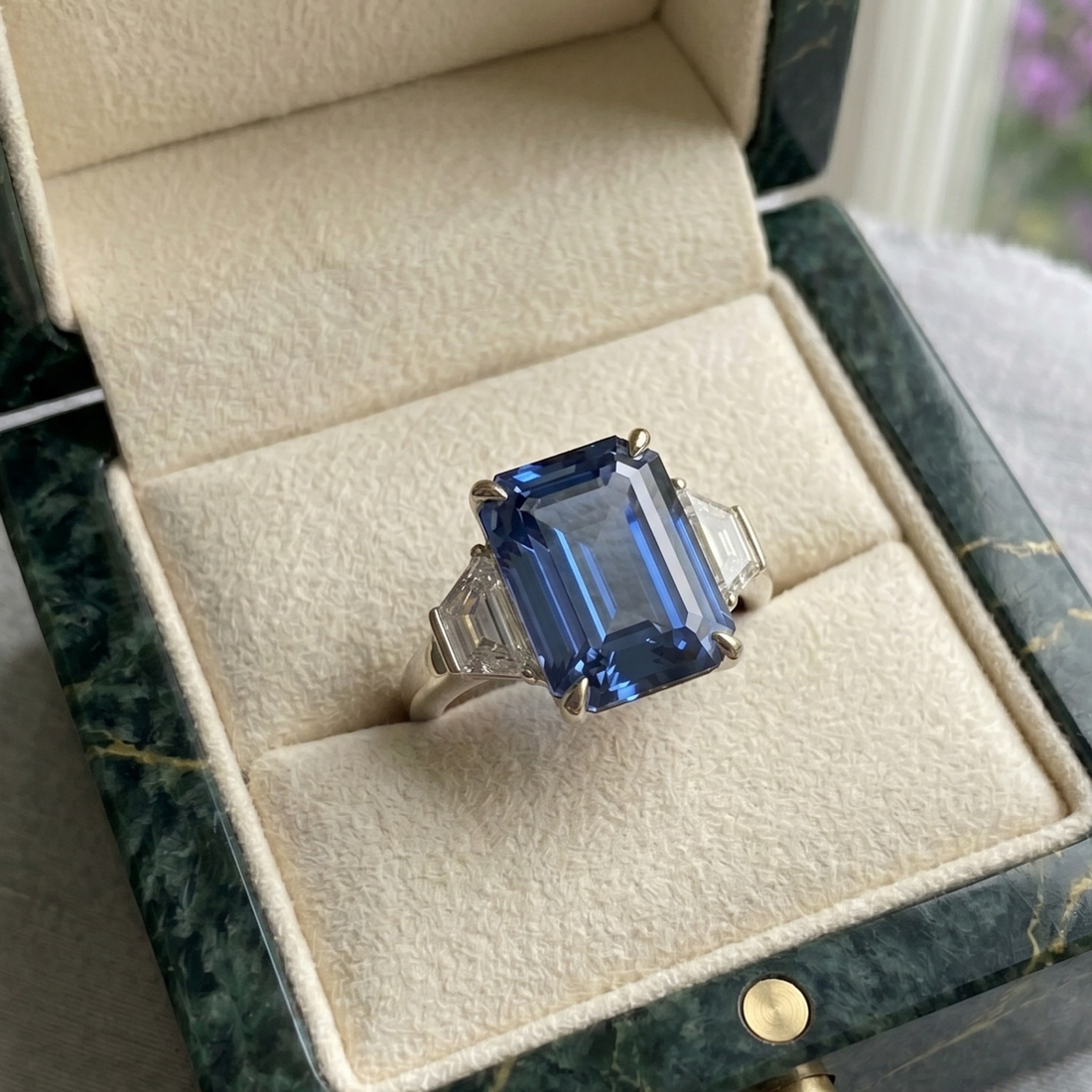 A silver ring with a large emerald-cut blue sapphire center stone and smaller side stones, displayed in an open cream velvet jewelry box with a dark green marble exterior.