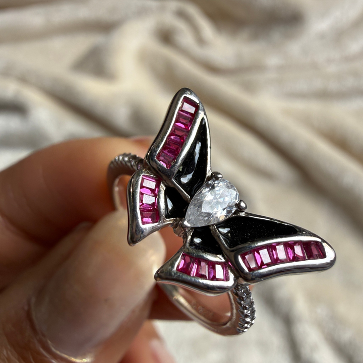 A butterfly-shaped ring with silver-toned metal, black enamel, and bright pink gemstone accents, held between fingers against a soft beige fabric background.