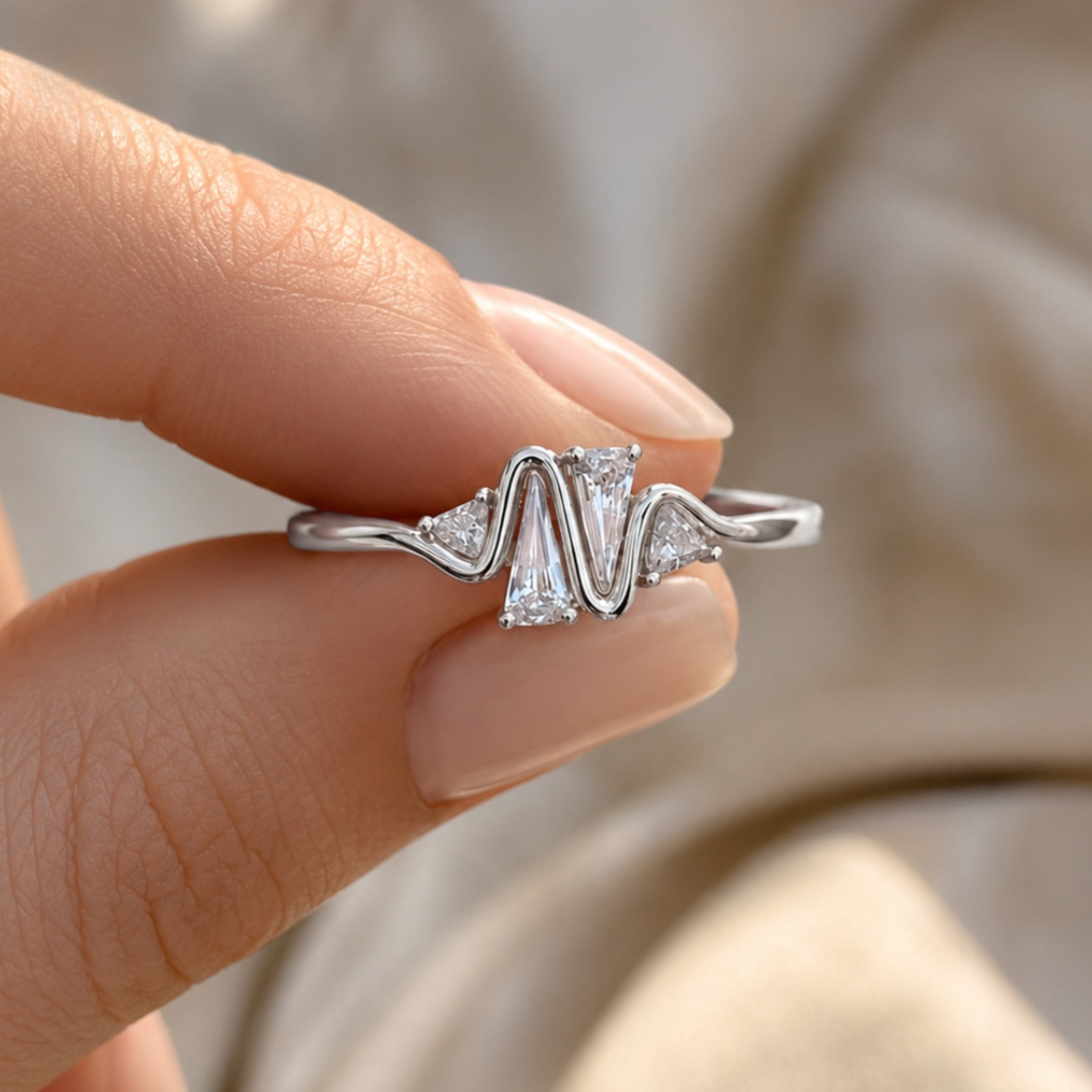 A silver ring with a wavy, zigzag-cut center and small clear accent stones, held between fingers against a soft beige fabric background.