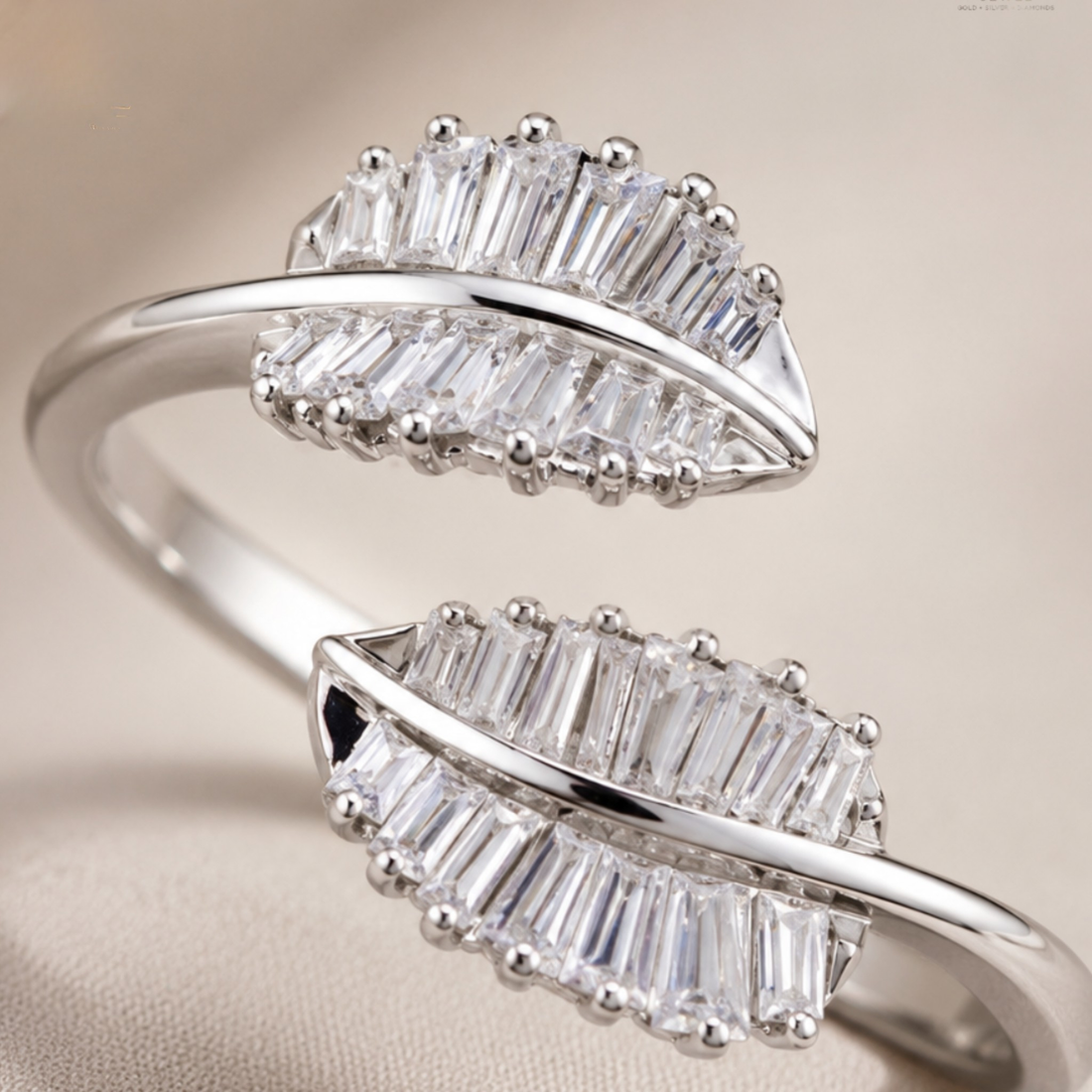 A silver-toned ring featuring two leaf-shaped clusters of clear baguette and round-cut stones, shown on a soft beige background.