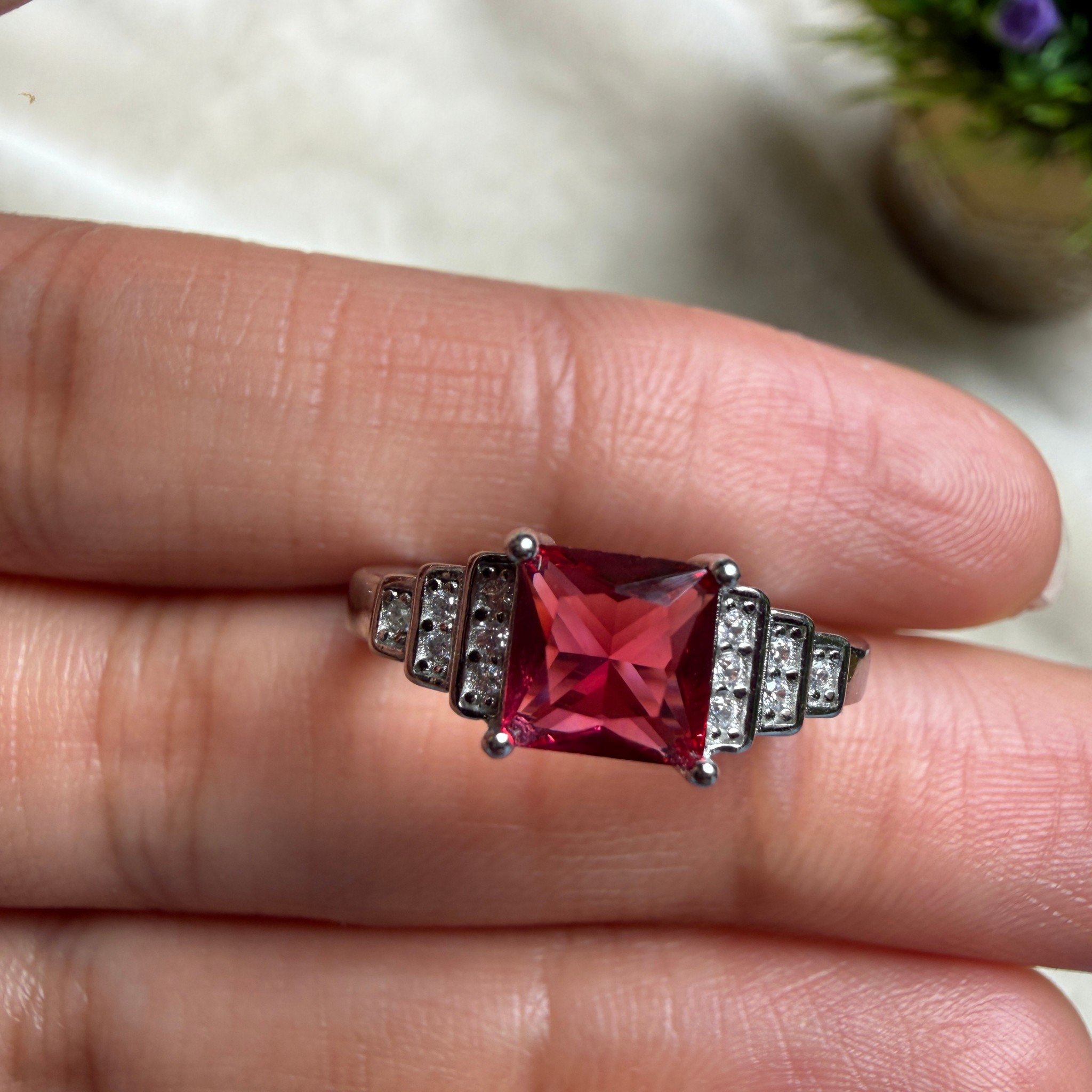 A silver ring with a large square pink gemstone and small clear stones on the shoulders, shown resting on a person's fingers.
