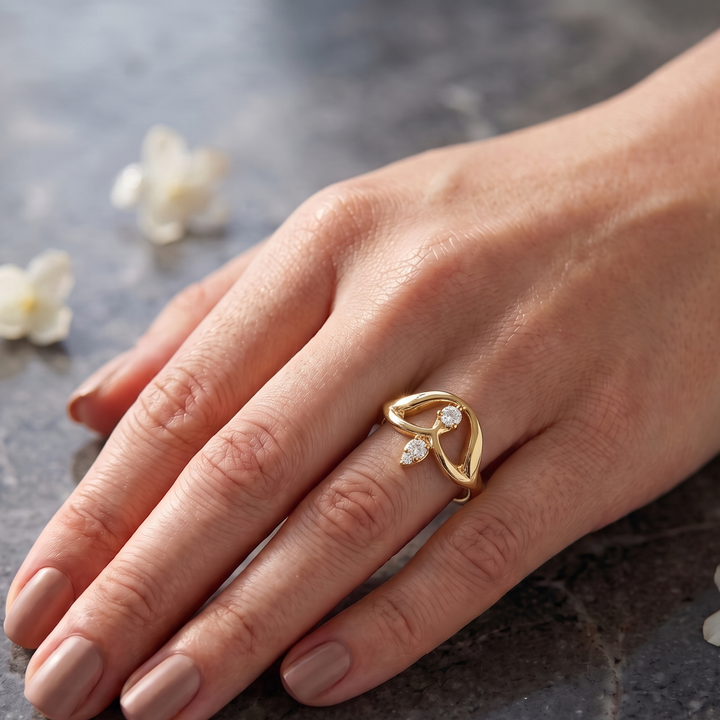 A gold ring with three small clear gemstones on a person's ring finger, shown against a dark textured background with blurred white flowers.