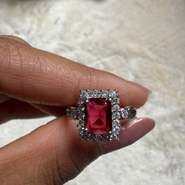 A silver ring with a rectangular red gemstone surrounded by small clear stones, held between fingers against a soft white textured background.
