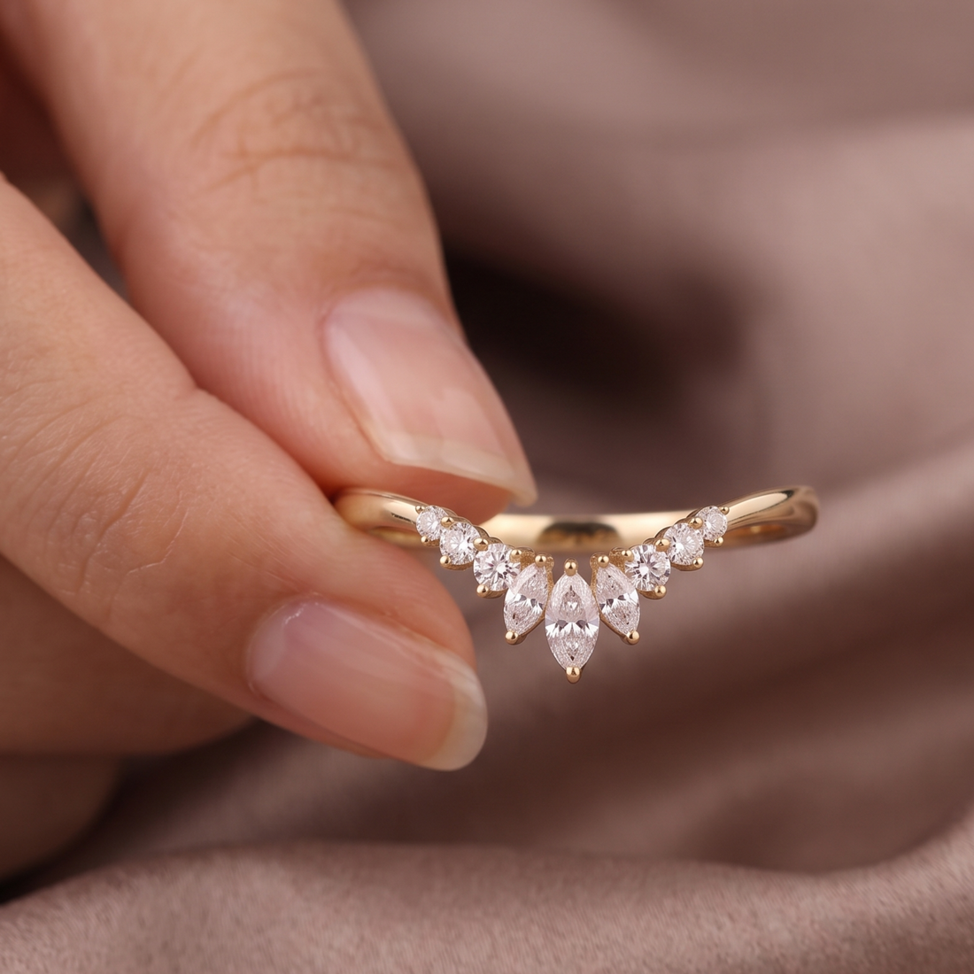 A close-up of a hand holding a delicate gold ring with a curved V-shaped setting adorned with small clear marquise and round stones, set against a soft brown fabric background.