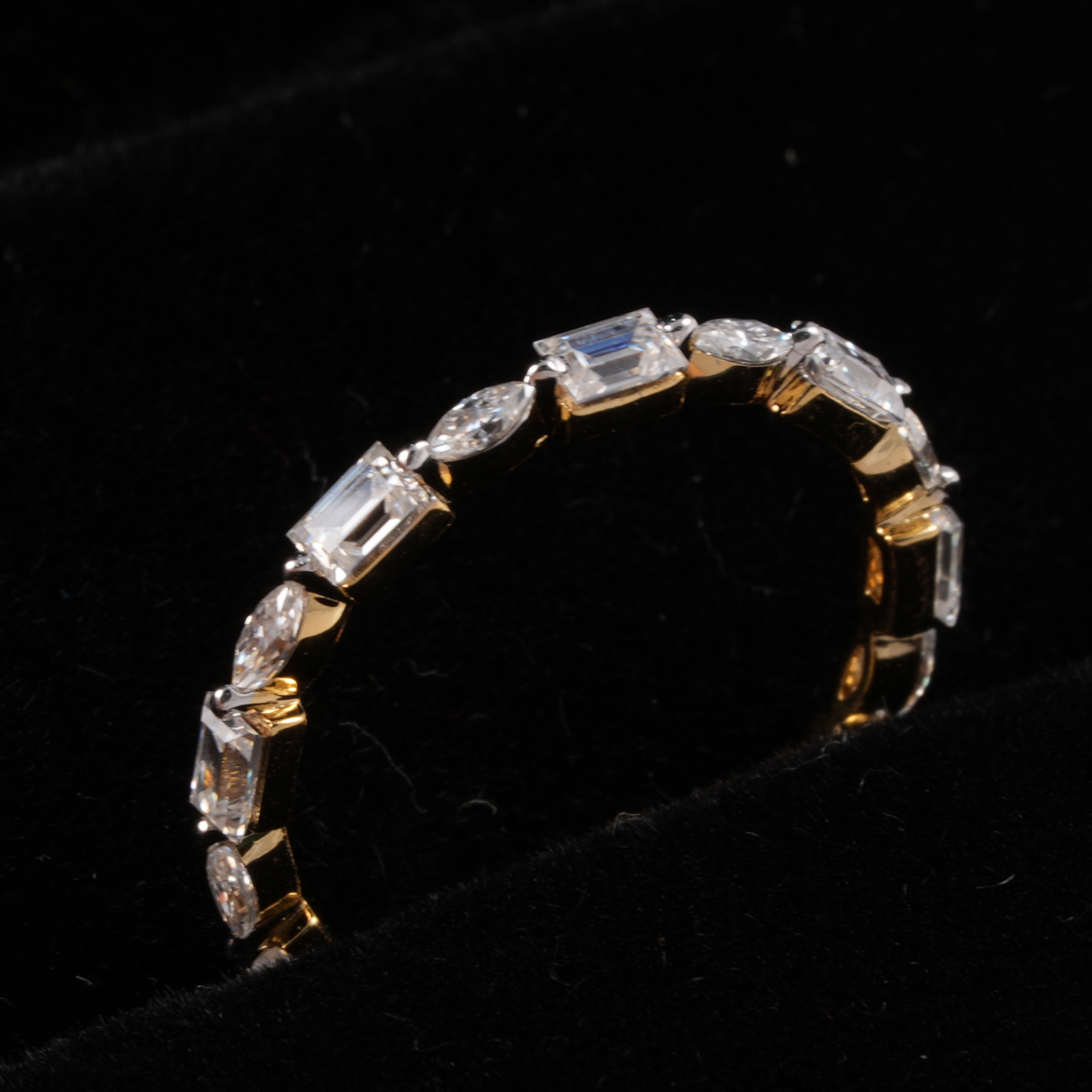 A gold-toned eternity ring featuring a row of clear rectangular and marquise-cut gemstones set against a black velvet background.