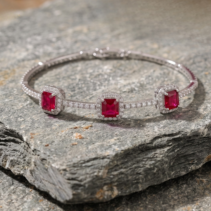 A silver-toned bangle bracelet with three square-cut red gemstones, each surrounded by a halo of small clear stones, displayed on a textured gray rock.