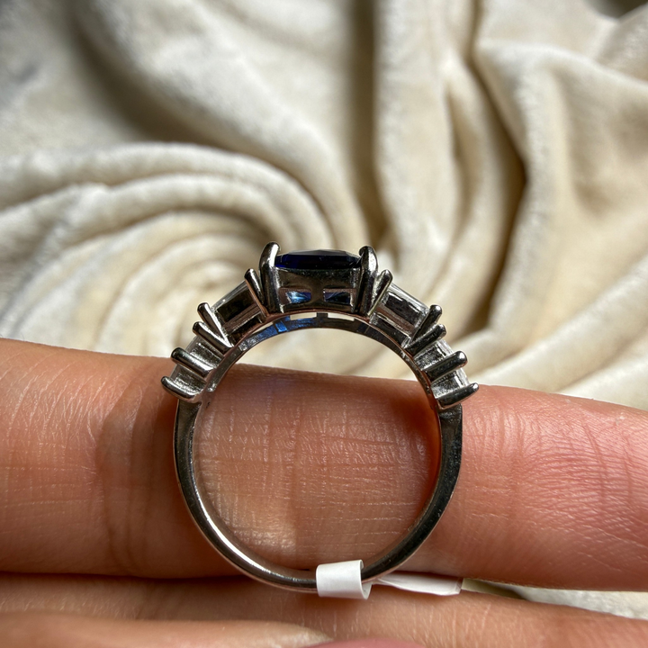 A silver-toned ring with a rectangular dark blue center stone and smaller side stones, shown resting on a person's fingers against a soft cream fabric background.