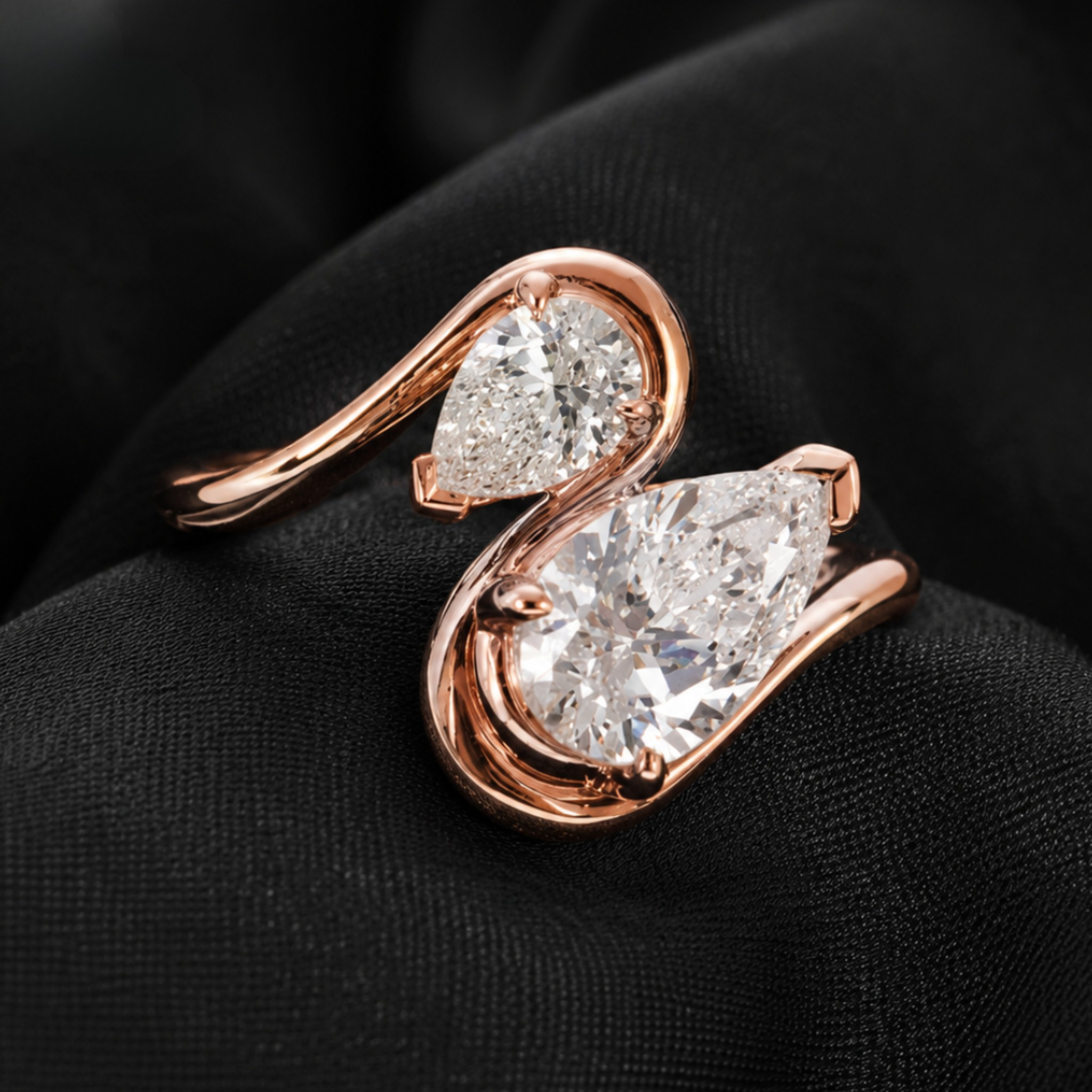 A rose gold ring featuring two pear-cut clear gemstones in a swan-like curved setting, displayed on a textured black fabric background.