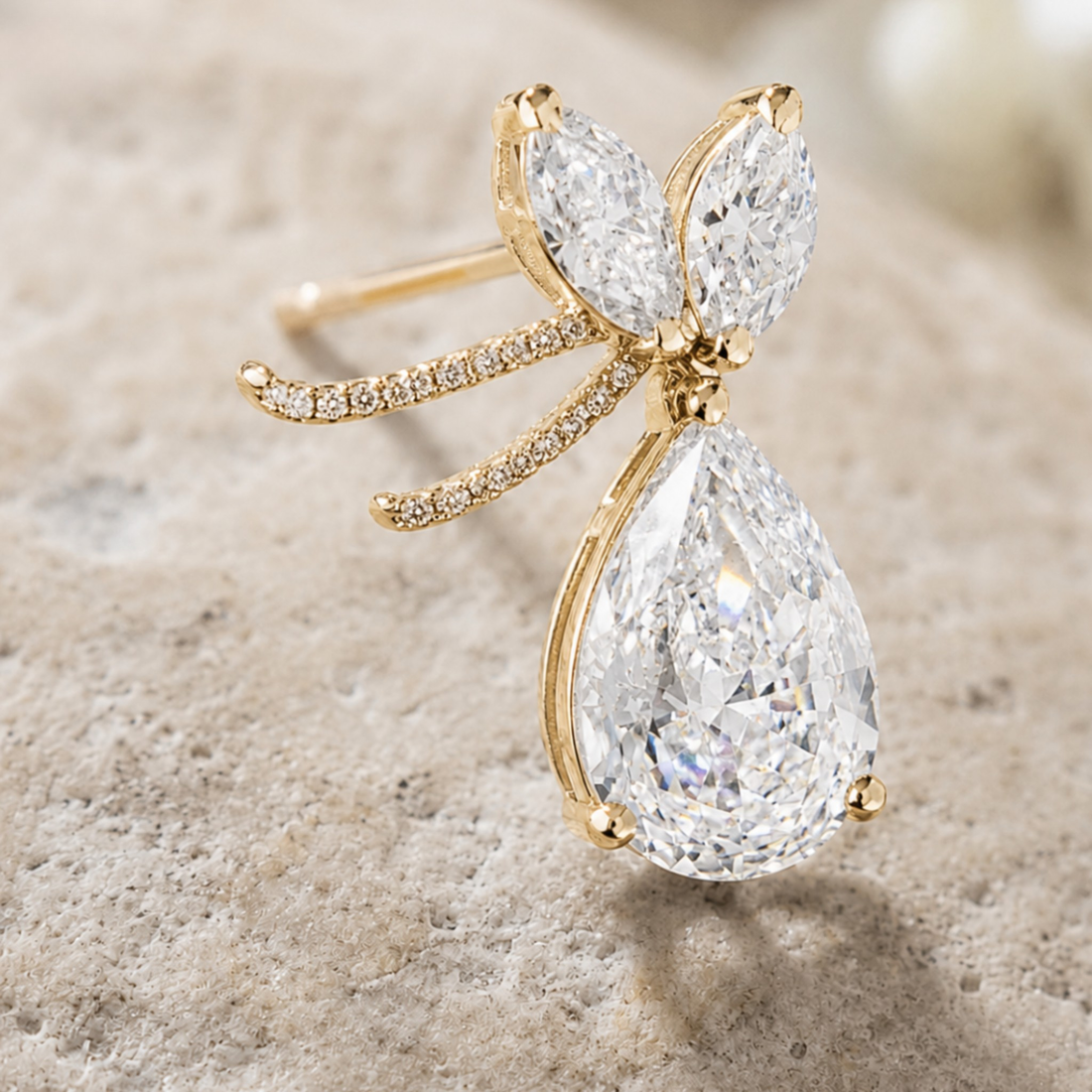 A gold butterfly ring featuring a large pear-cut clear gemstone and smaller marquise-cut stones, accented with tiny pavé diamonds on the wings and band, set against a textured light beige stone background.