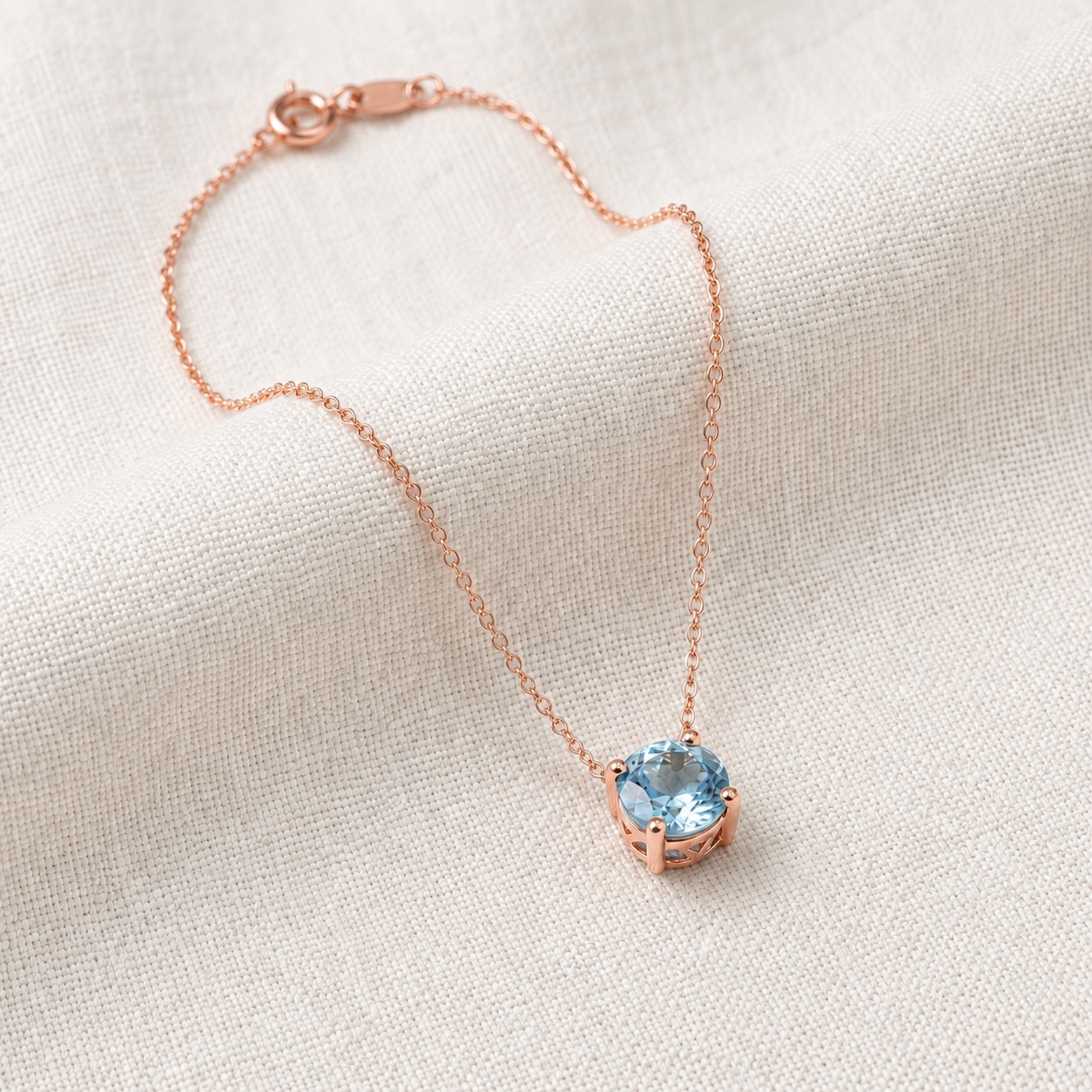 A delicate rose gold chain necklace with a small round bezel-set light blue gemstone pendant, displayed on a textured off-white linen fabric background.
