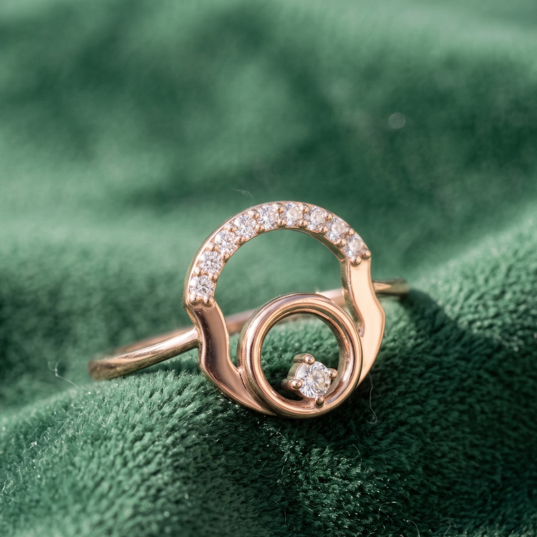 A rose gold ring with a double-ring design, accented by small clear pavé stones on the top arch and a single round stone on the lower right, resting on a textured dark green velvet surface.