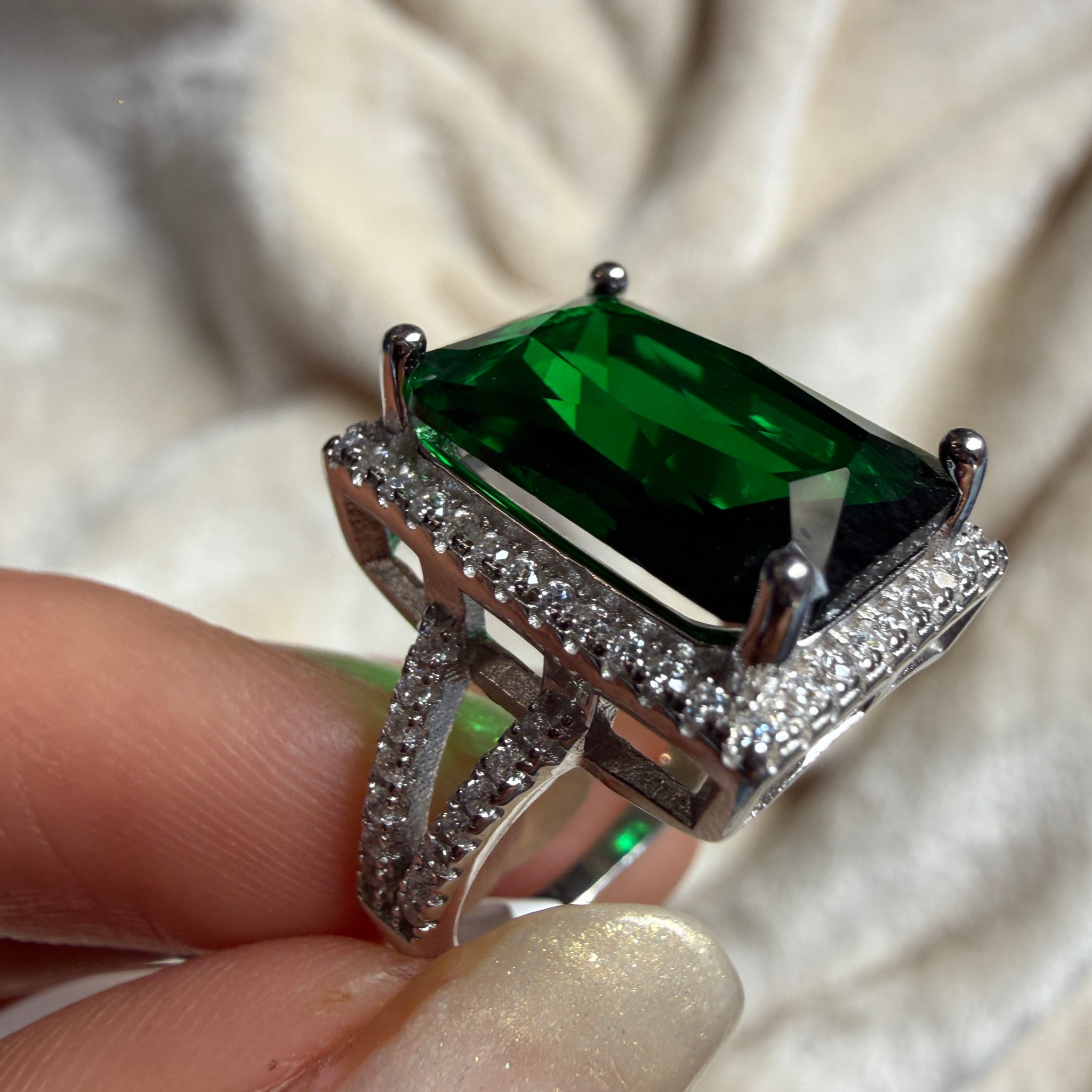 A close-up of a silver ring with a large rectangular green emerald-cut gemstone and pavé-set clear stones, held between fingers against a soft cream fabric background.