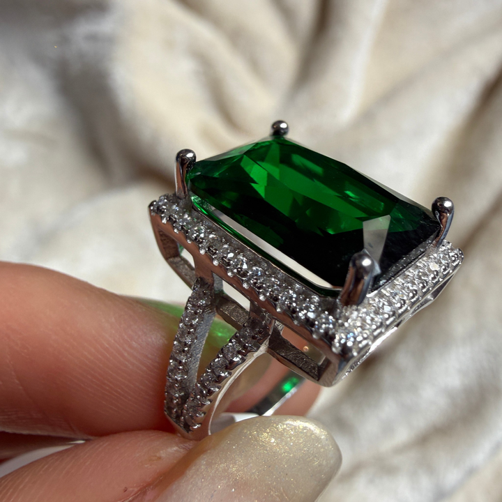 A close-up of a silver ring with a large rectangular green emerald-cut gemstone and pavé-set clear stones, held between fingers against a soft cream fabric background.