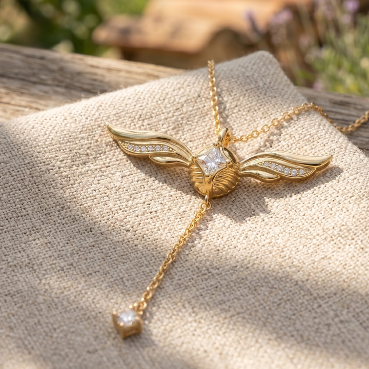 A gold-toned pendant necklace with a winged design and a dangling chain ending in a small clear gemstone, displayed on a textured beige fabric surface outdoors.