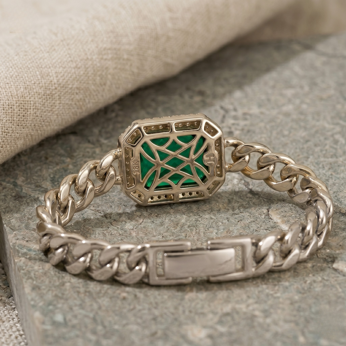 A silver-toned chunky chain bracelet with a geometric octagonal charm featuring a green starburst pattern, displayed on a textured gray stone surface with a beige linen cloth in the background.