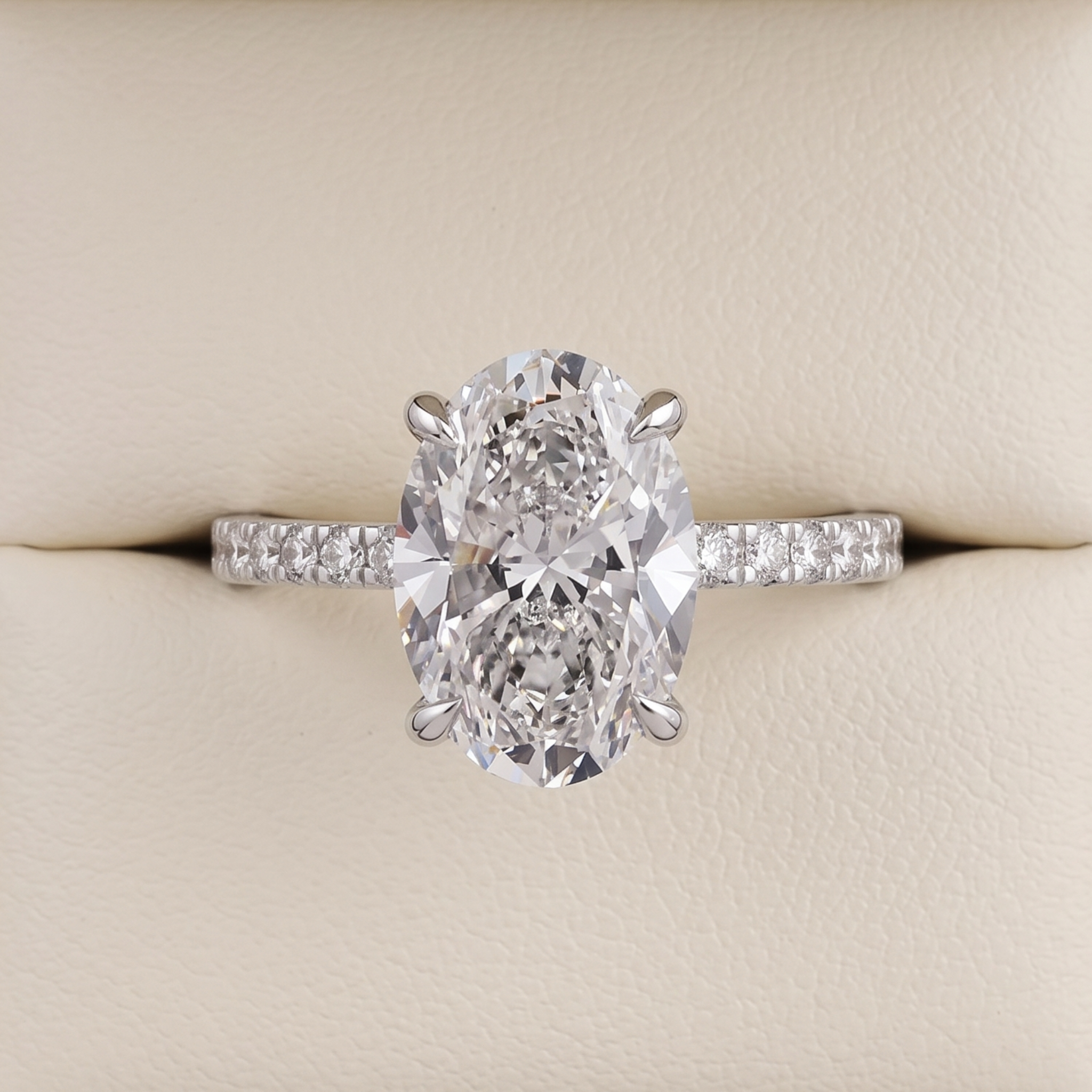 An oval-cut diamond engagement ring with a silver-colored band set with small diamonds, displayed on a cream-colored ring box.