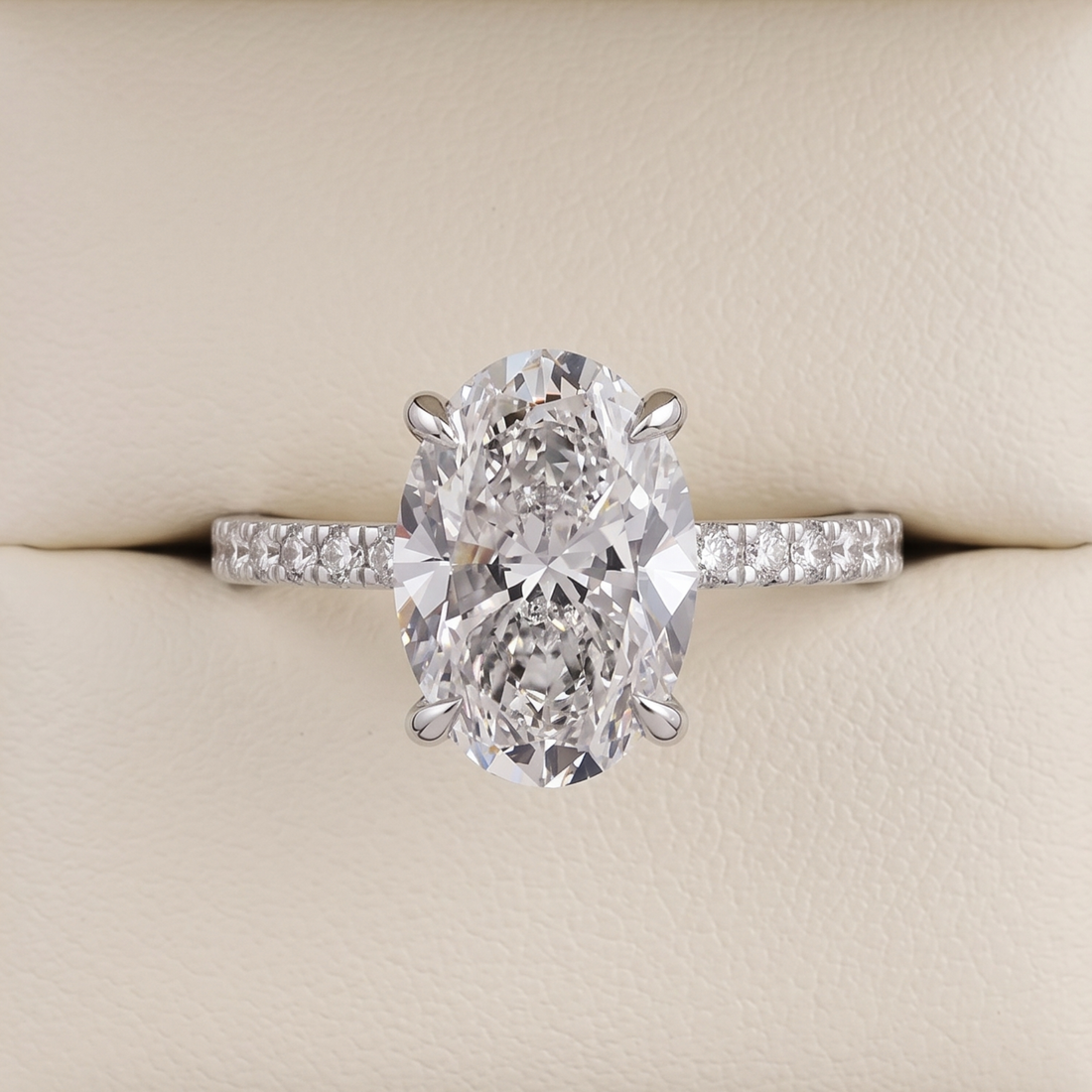 An oval-cut diamond engagement ring with a silver-colored band set with small diamonds, displayed on a cream-colored ring box.