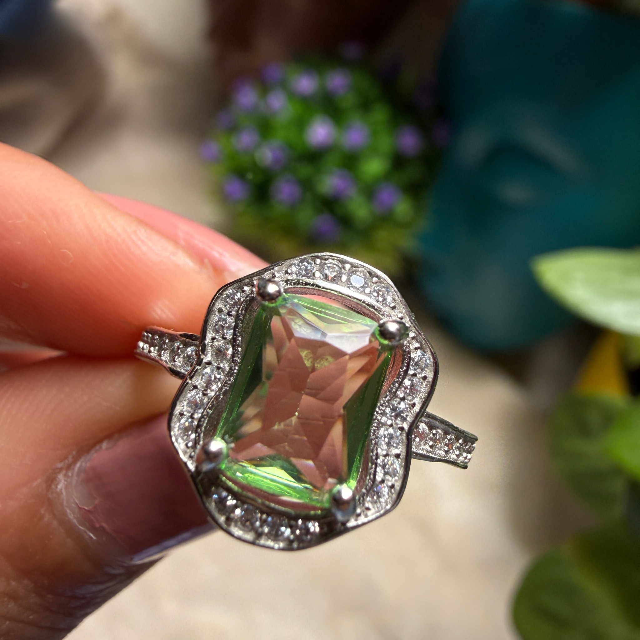 A close-up of a hand holding a silver ring featuring a large iridescent green and pink cushion-cut gemstone surrounded by a halo of small clear stones.