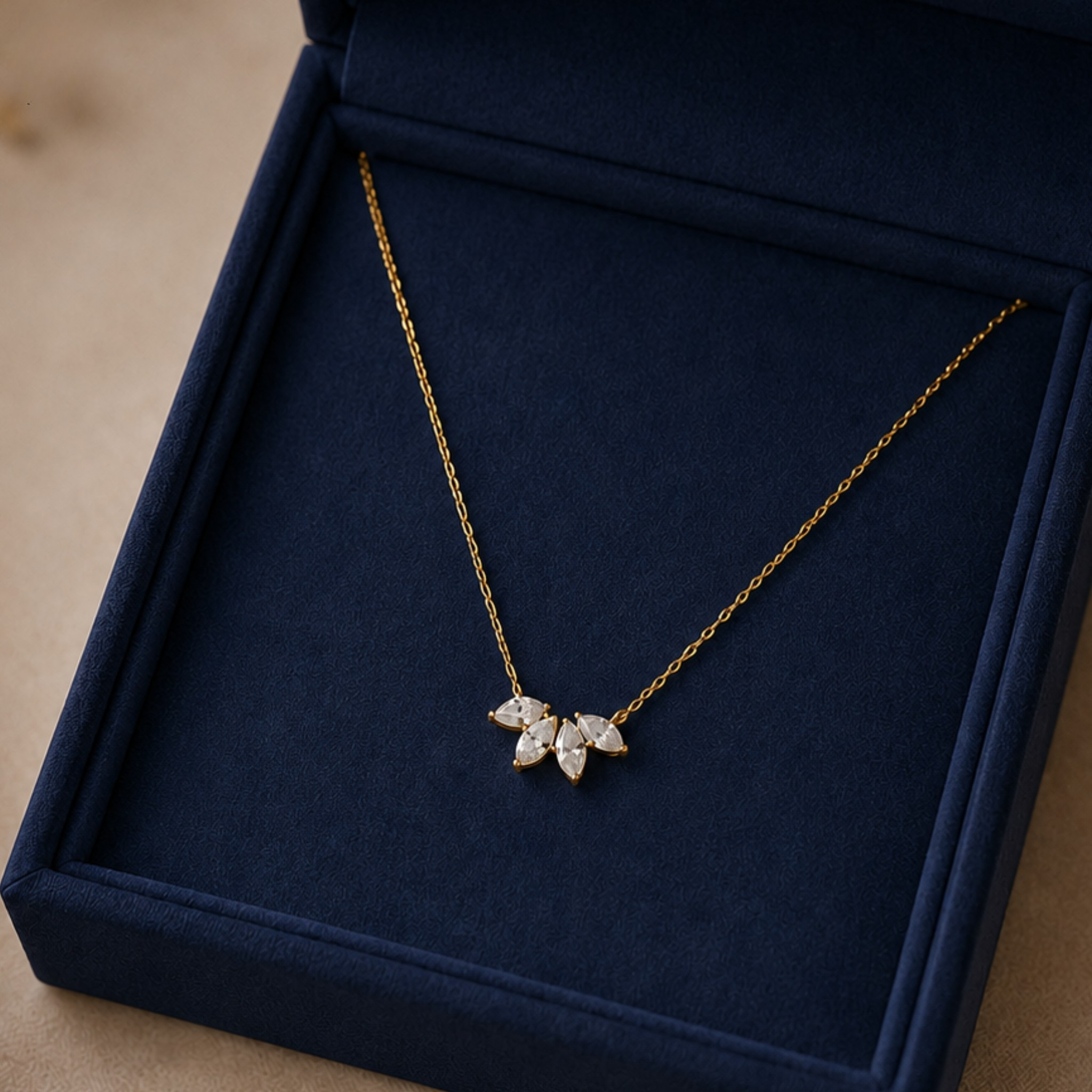 A delicate gold chain necklace with a small leaf-like pendant set with clear stones, displayed inside an open dark navy velvet jewelry box.