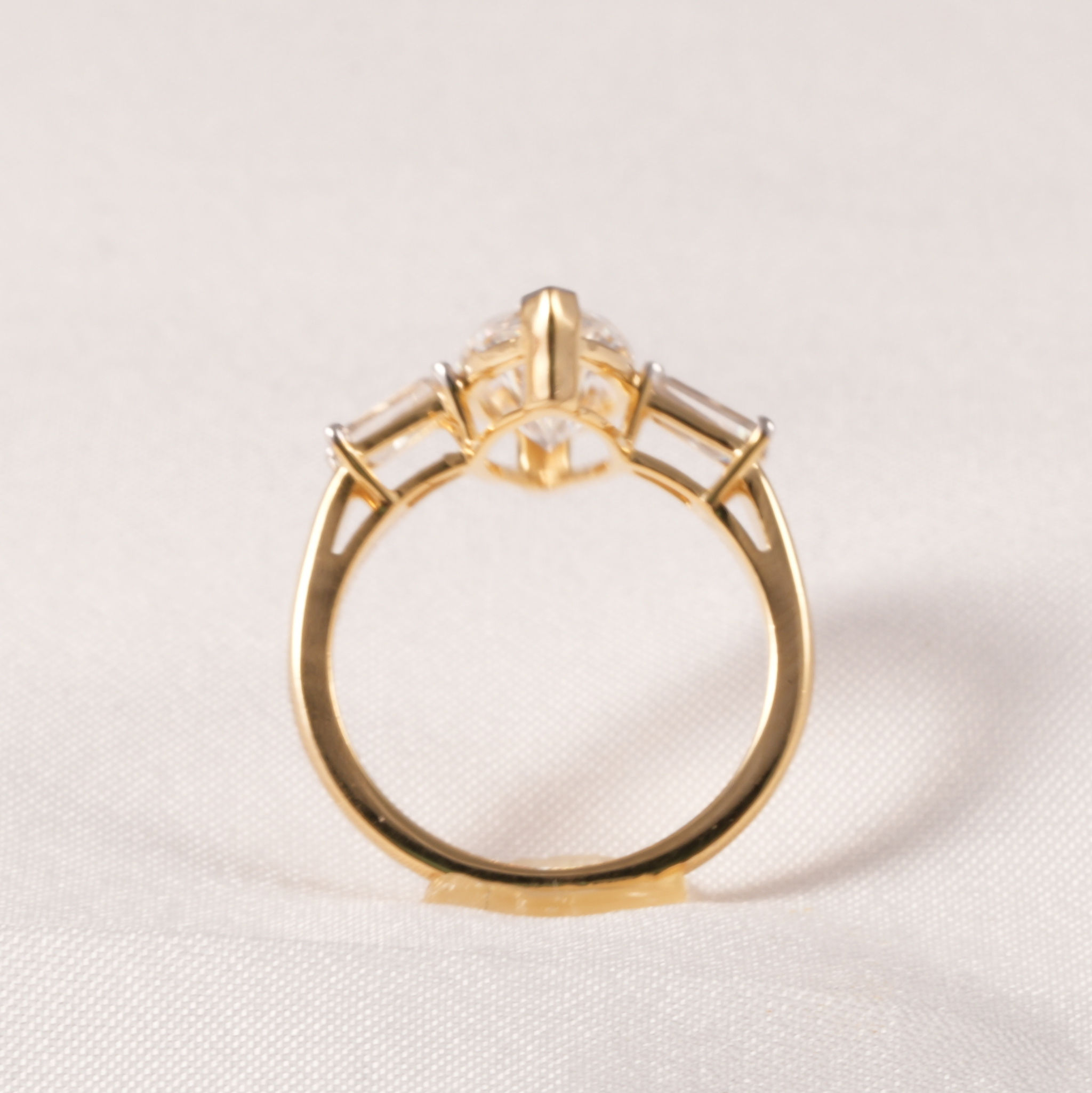 A yellow gold ring with a central round clear gemstone and two smaller rectangular side stones, photographed on a light textured fabric background.