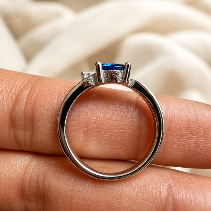 A silver ring with a rectangular blue gemstone in a prong setting, held on a person's fingers against a soft cream-colored fabric background.