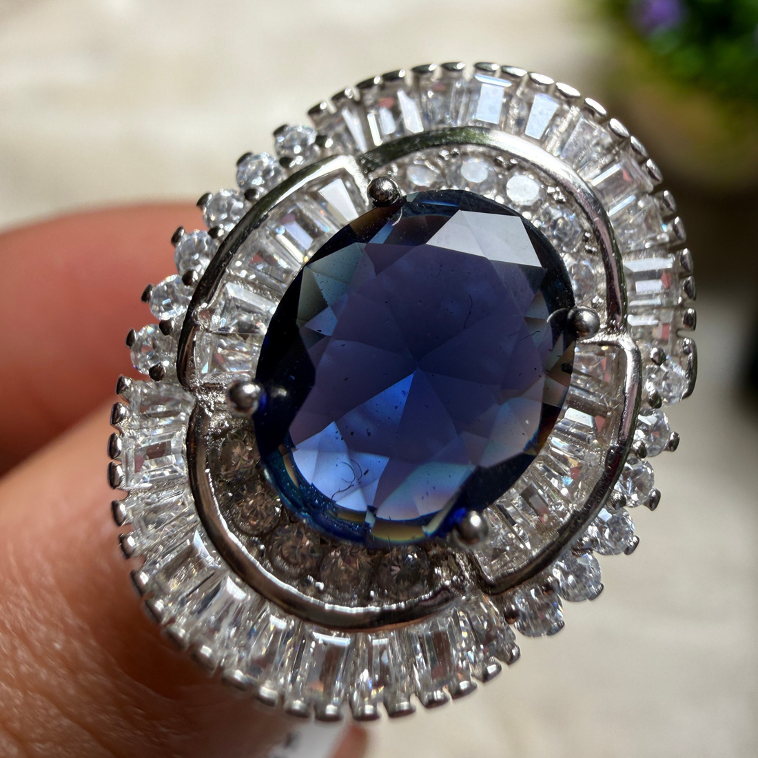 A close-up of a silver-toned ring with a large oval deep blue gemstone and surrounding clear baguette and round stones, shown being worn on a finger.