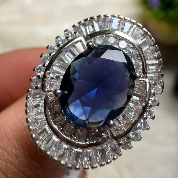 A close-up of a silver-toned ring with a large oval deep blue gemstone and surrounding clear baguette and round stones, shown being worn on a finger.