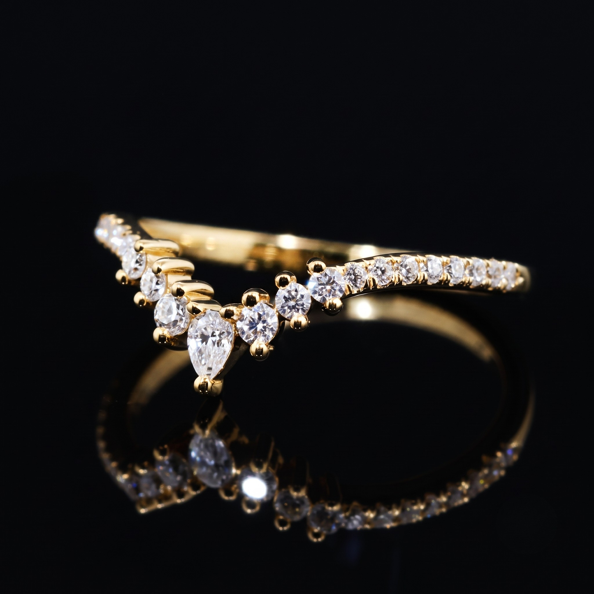 A yellow gold ring featuring a curved row of small round-cut clear stones and a central pear-cut clear stone, displayed on a reflective black surface against a black background.