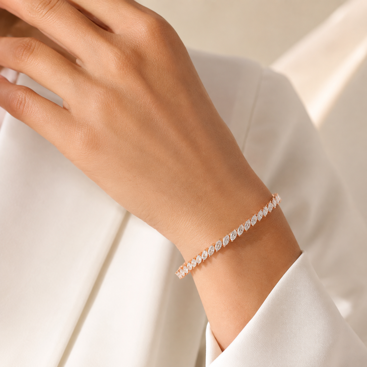 A close-up of a person's wrist wearing a delicate rose gold tennis bracelet with small clear stones, styled with a white blazer.