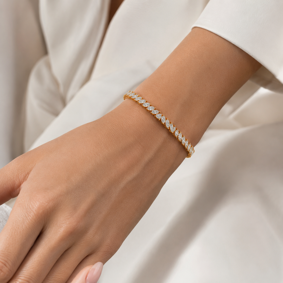 A delicate gold tennis bracelet with small clear stones, worn on a woman's wrist against a soft white dress.