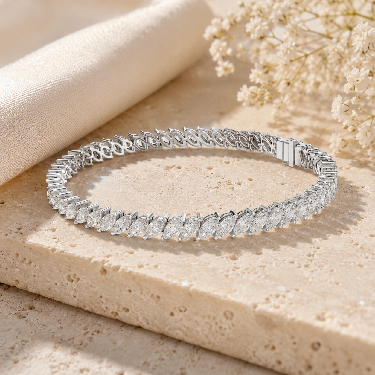 A silver tennis bracelet with marquise-cut stones, displayed on a textured beige stone slab with a cream fabric napkin and dried baby's breath flowers.