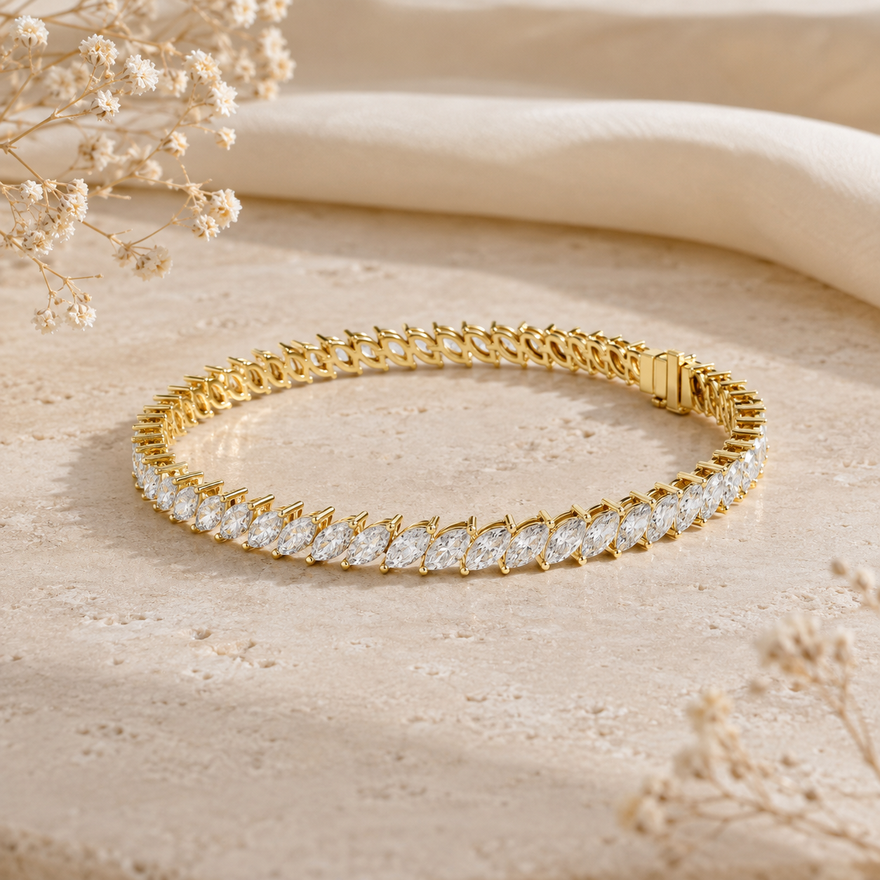 A gold tennis bracelet with clear marquise-cut stones, displayed on a textured beige surface with dried flowers and a soft cream fabric backdrop.