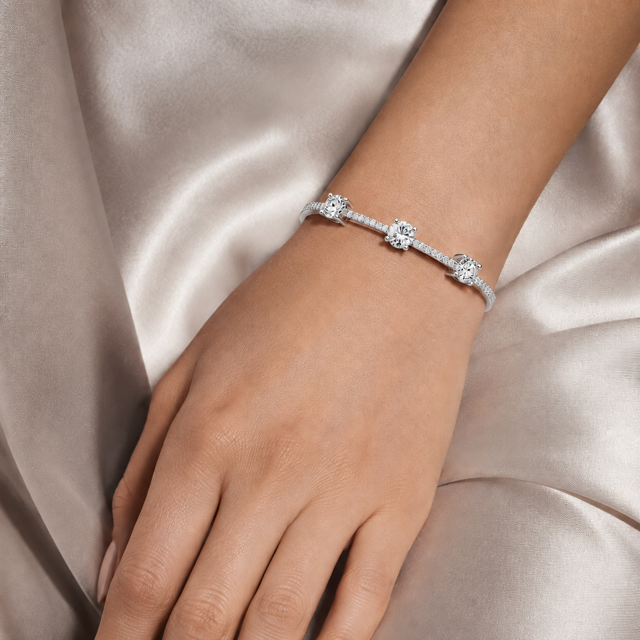 A close-up of a person's wrist wearing a delicate silver-toned bracelet with three round-cut diamond clusters, set against a soft champagne satin fabric background.