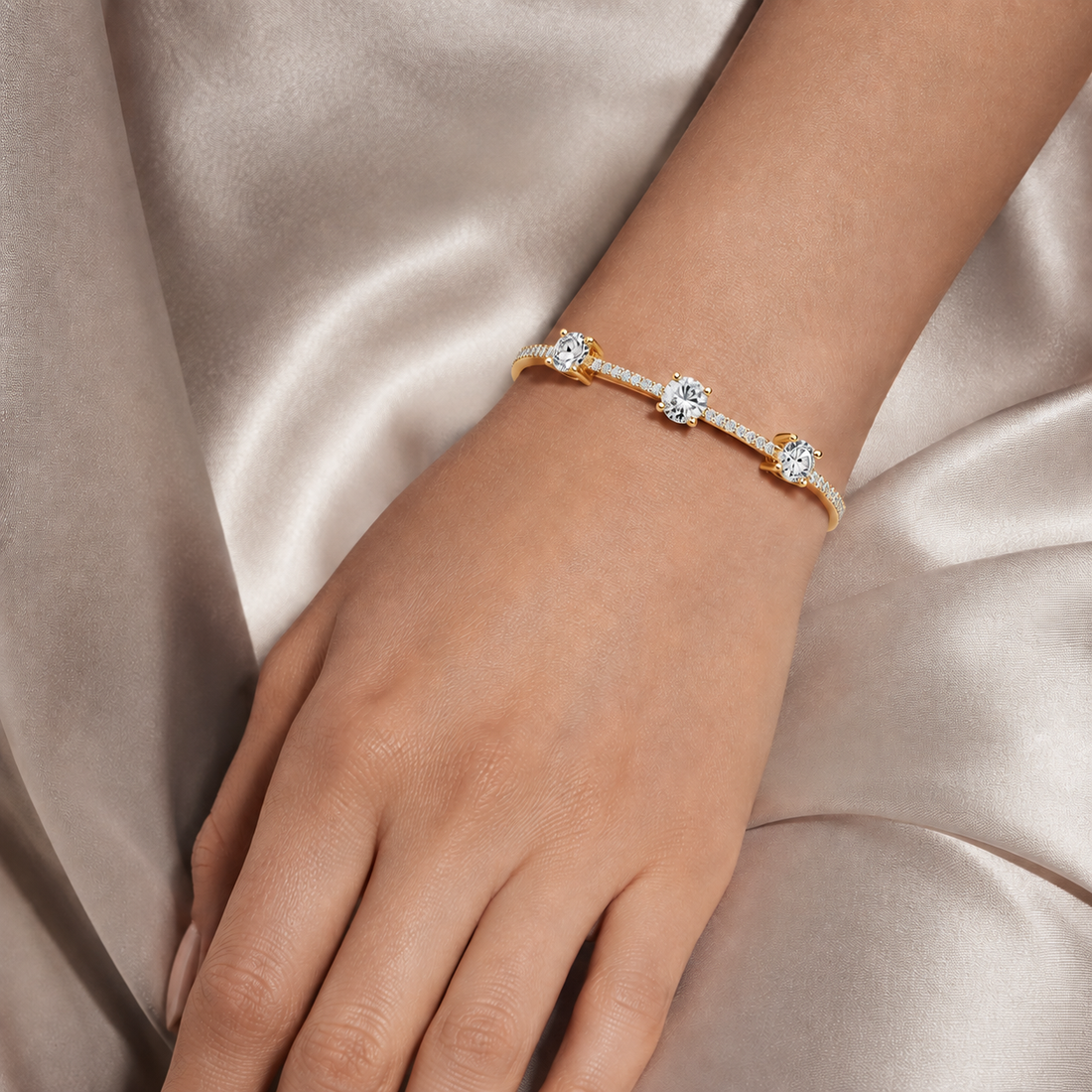 A delicate gold bracelet with three round-cut clear gemstones and small pavé accents, shown worn on a hand against a satin fabric background.