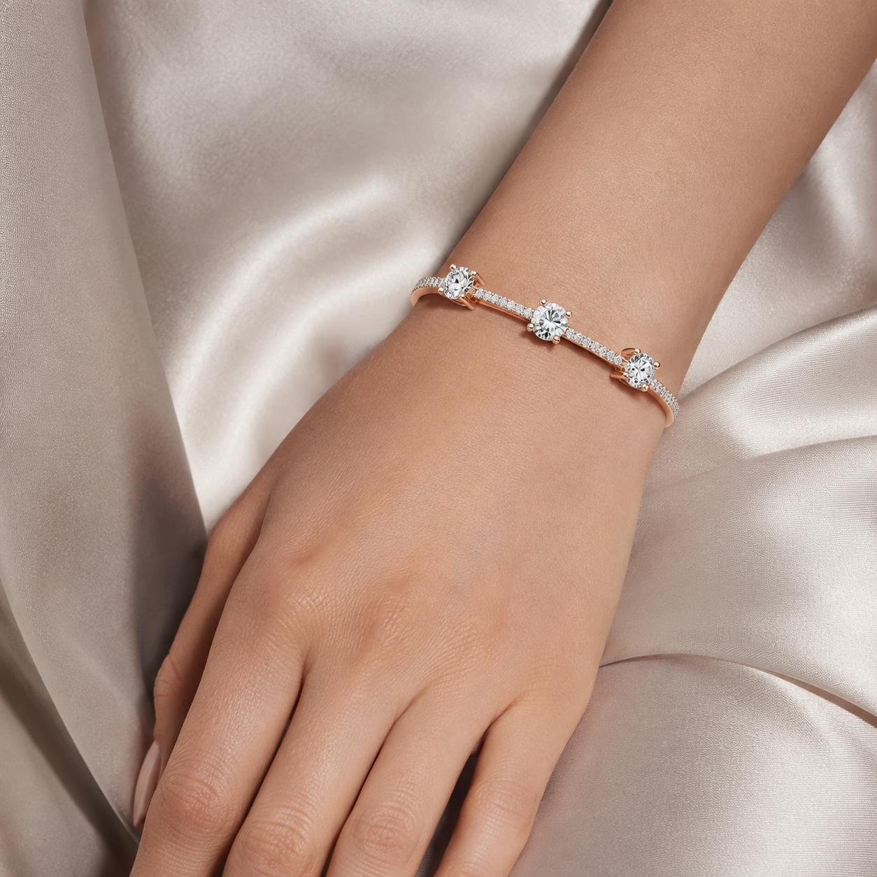 A delicate bracelet with three round-cut clear stones on a rose gold band, shown worn on a woman's wrist against a soft champagne satin background.