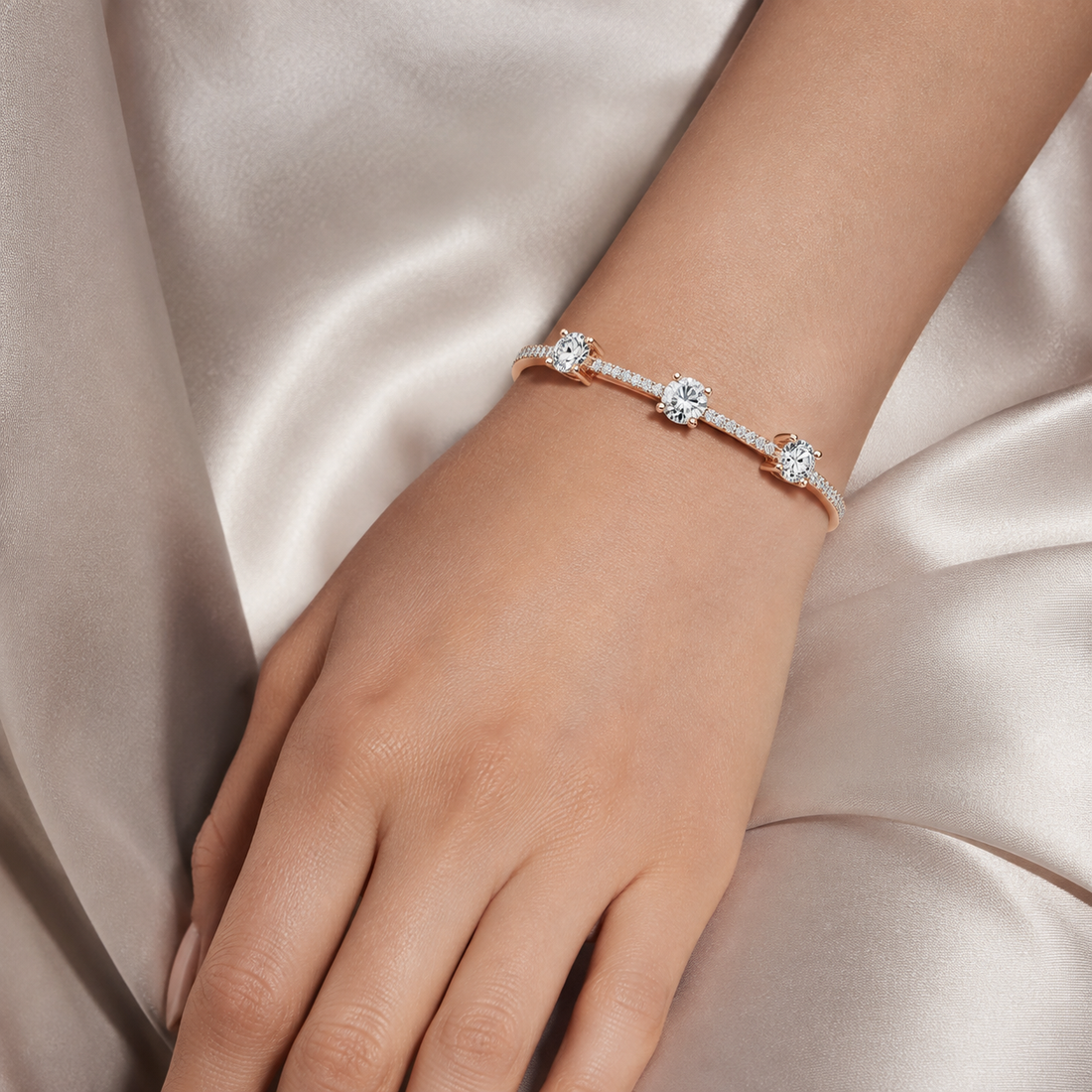 A delicate bracelet with three round-cut clear stones on a rose gold band, shown worn on a woman's wrist against a soft champagne satin background.