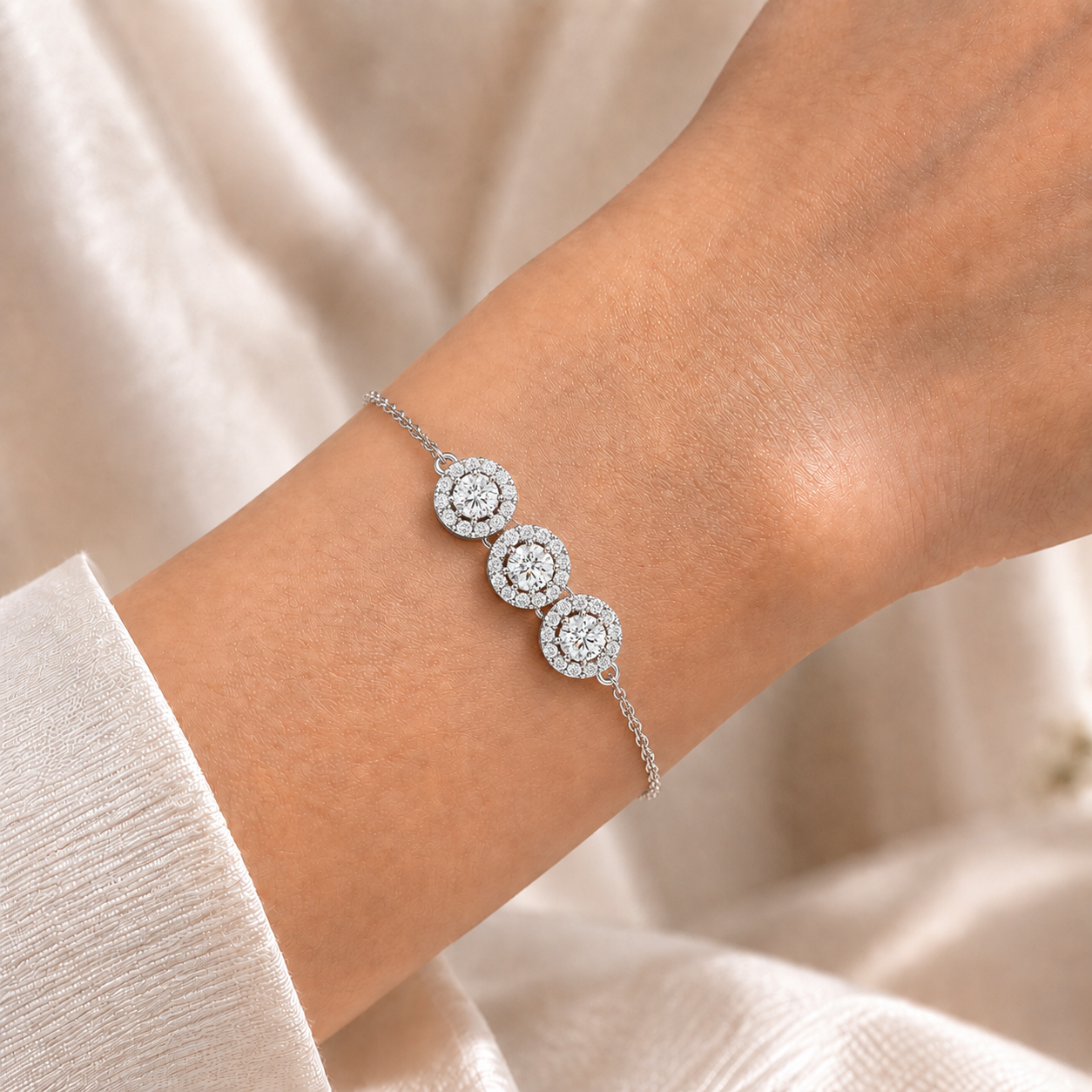 A delicate silver chain bracelet with three circular cluster motifs set with clear stones, shown worn on a person's wrist against a soft beige fabric background.