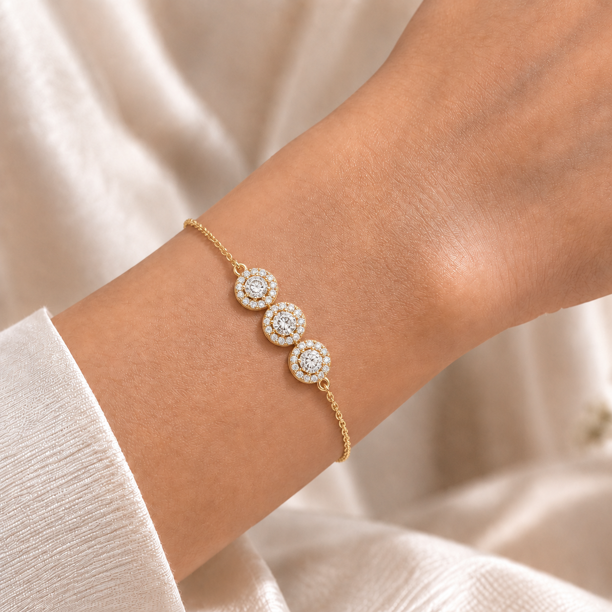 A delicate gold chain bracelet with three circular cluster motifs set with clear stones, shown worn on a person's wrist against a soft cream fabric background.