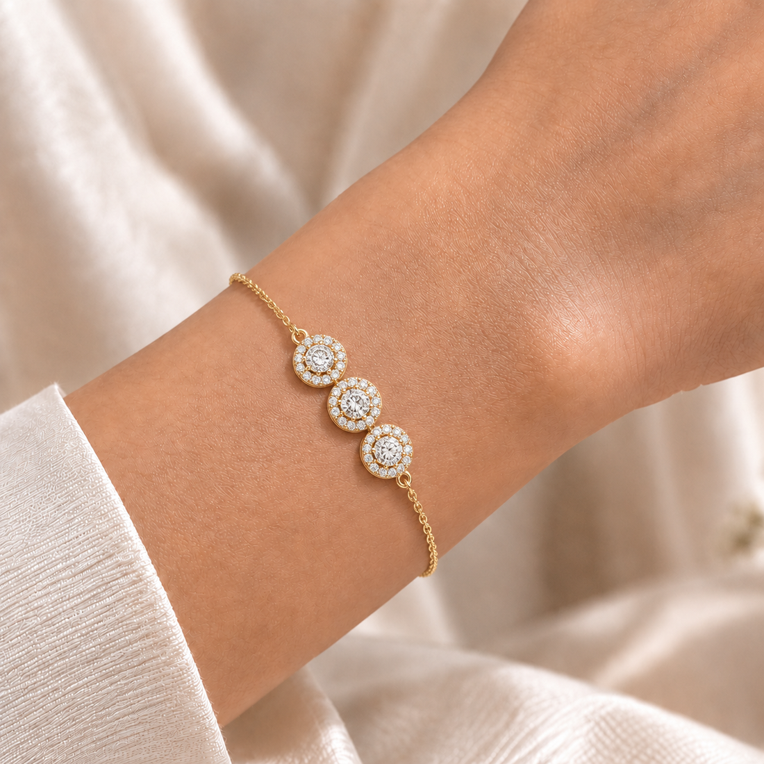 A delicate gold chain bracelet with three circular cluster motifs set with clear stones, shown worn on a person's wrist against a soft cream fabric background.