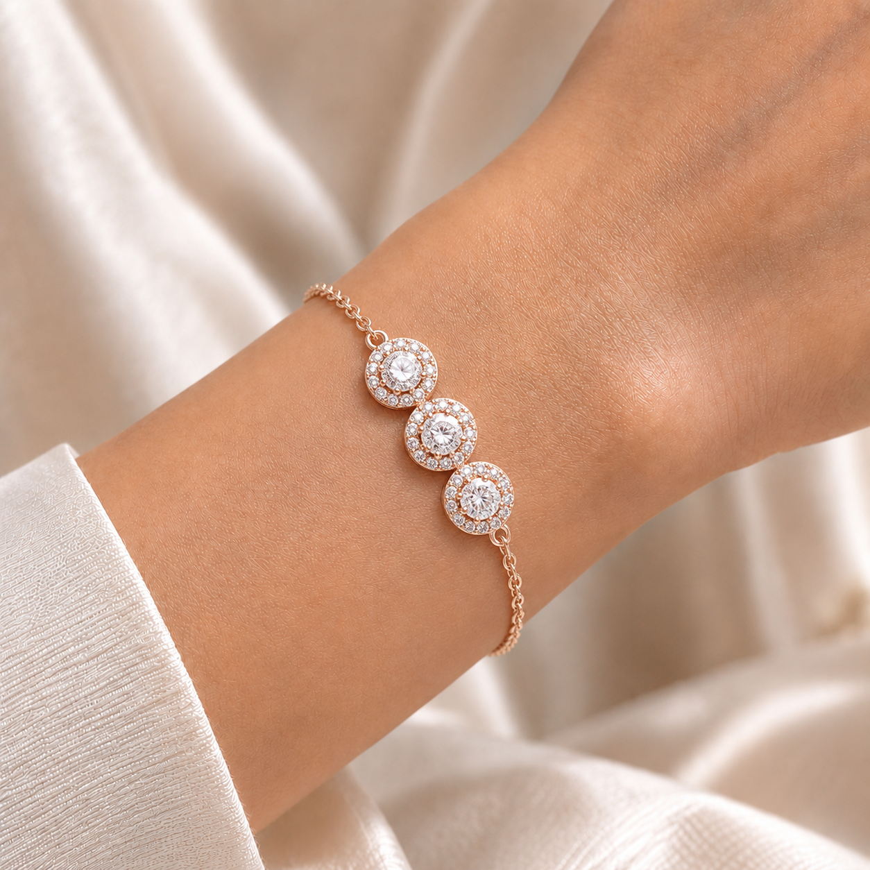 A close-up of a delicate rose gold chain bracelet with three circular pavé diamond clusters worn on a wrist, set against a soft, draped cream-colored fabric background.