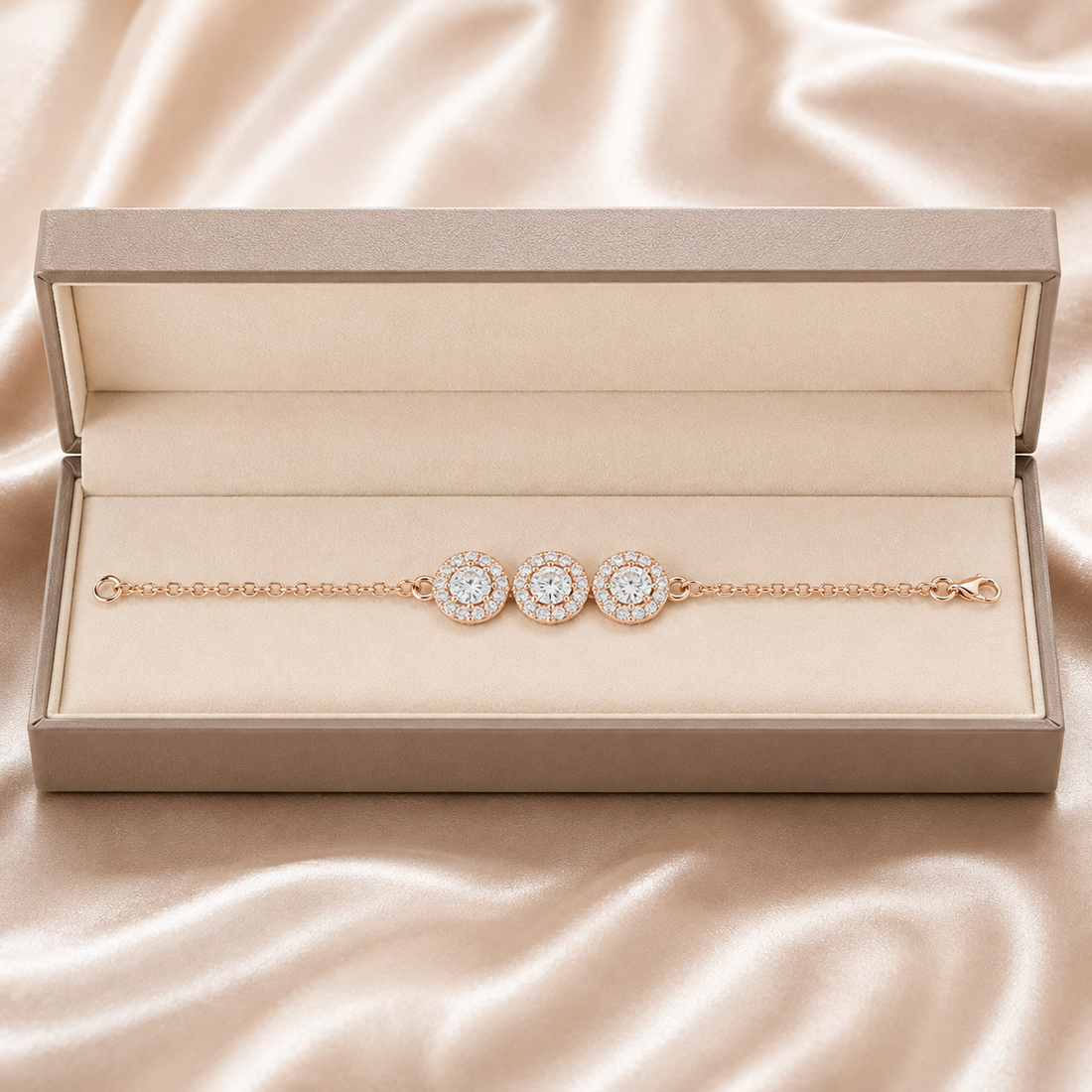 A rose gold chain bracelet with three round halo diamond pendants, presented in an open champagne-colored jewelry box on a draped satin fabric background.