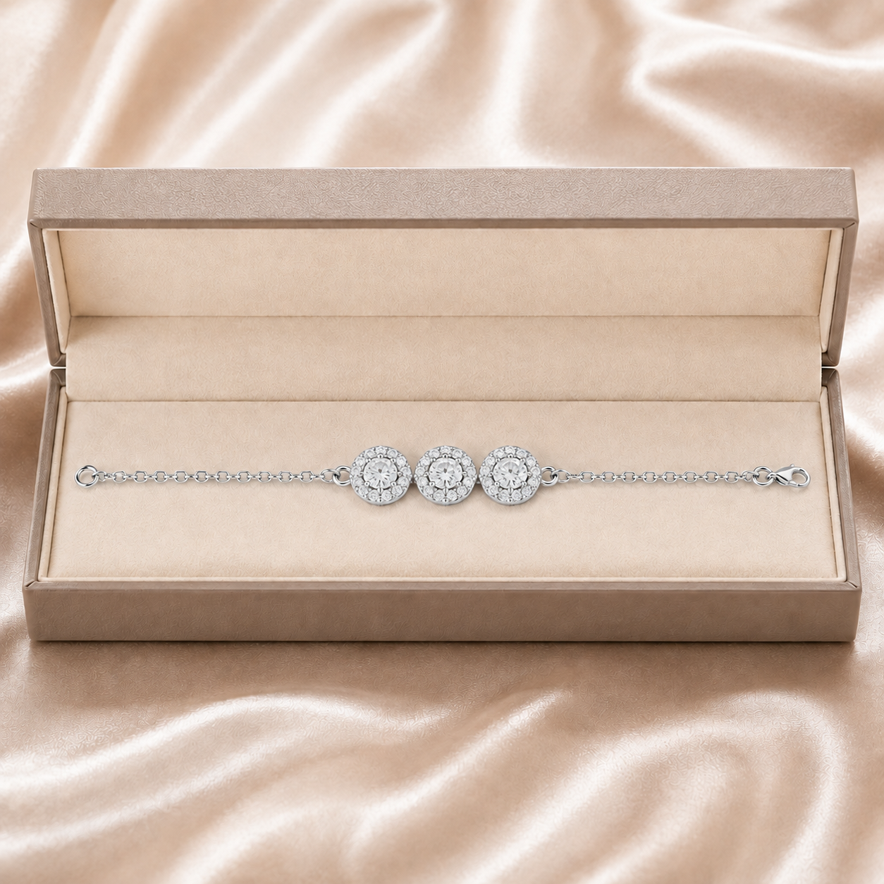 A delicate silver-toned chain bracelet with three round halo diamond motifs, presented in an open beige jewelry box on a satin fabric background.
