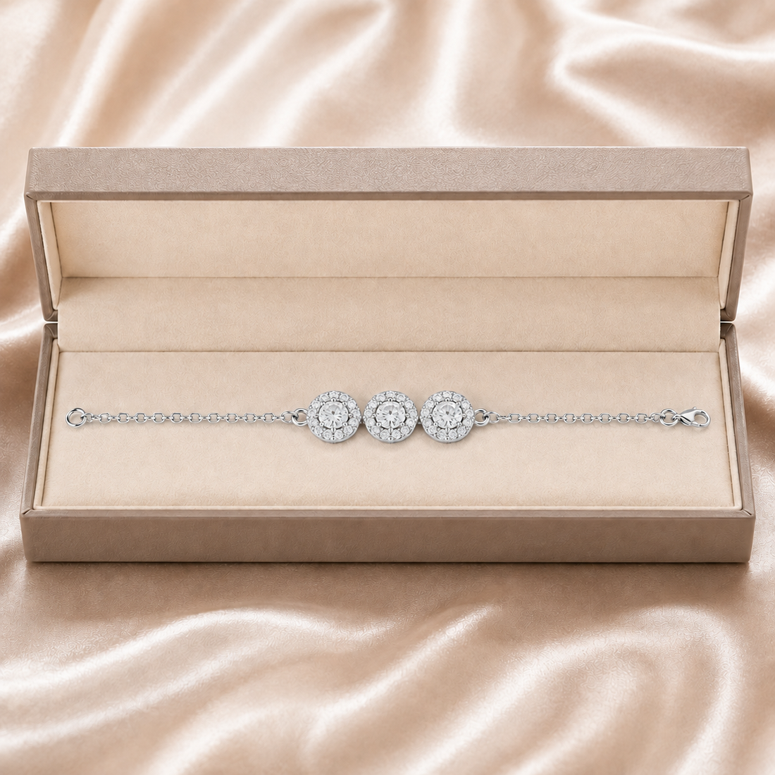 A delicate silver-toned chain bracelet with three round halo diamond motifs, presented in an open beige jewelry box on a satin fabric background.